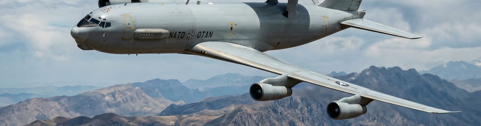 NATO E-3A Sentry AWACS aircraft in flight over mountainous terrain showing distinctive radar dome above fuselage